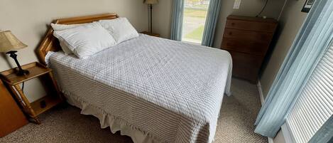 Una cama bien hecha con colcha a rayas y almohadas, una lámpara de noche, una mesita de madera, un aparador, un televisor montado en la pared y una ventana con cortinas.