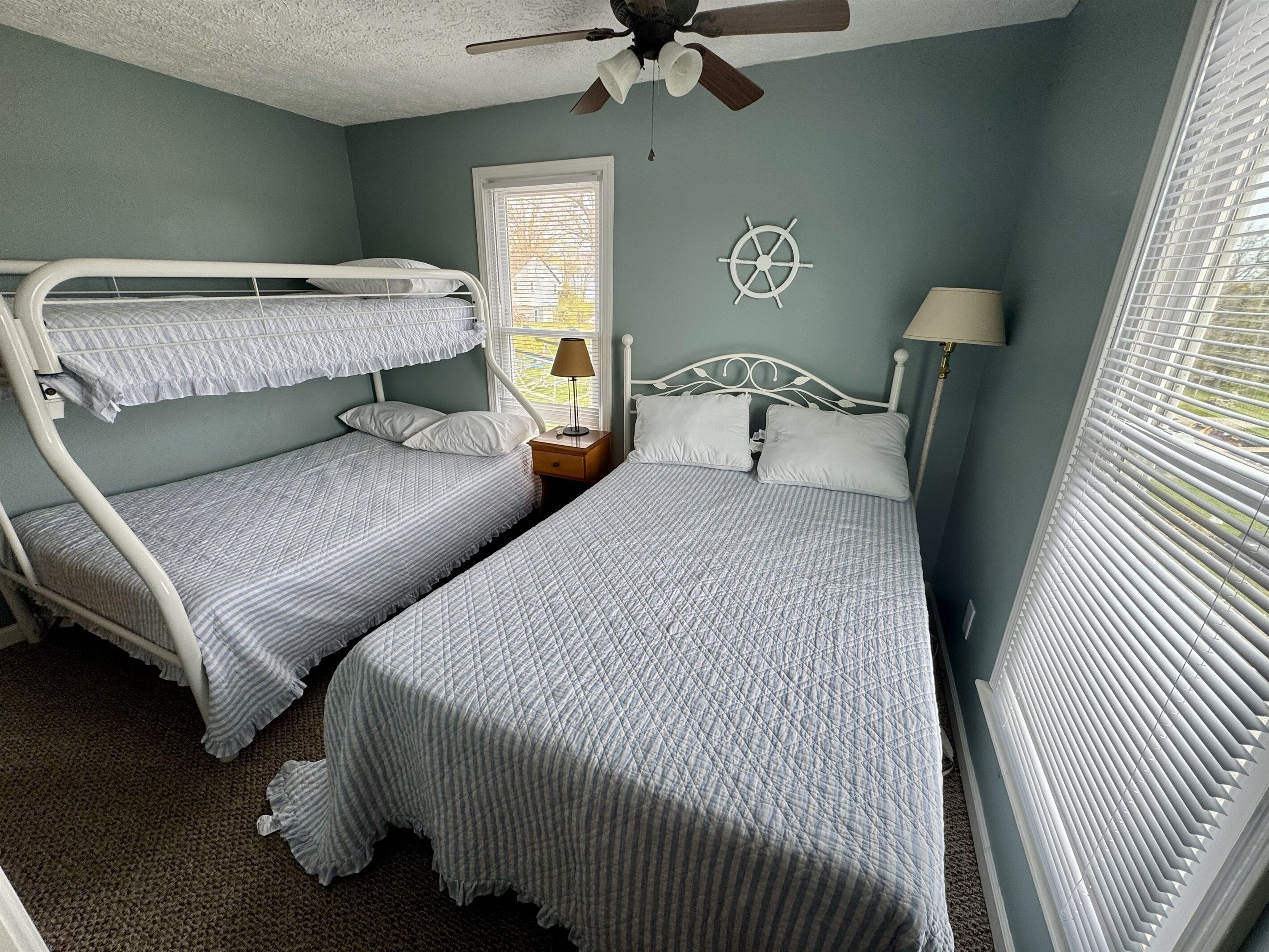 Une chambre avec des lits superposés, un ventilateur de plafond, une décoration en forme de roue de navire et une fenêtre avec des stores.