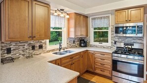 A modern kitchen with wooden cabinets, a stainless steel stove, and a large window with a view of greenery. - Sodus Bay View at the Point (Sodus Point)