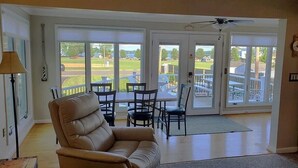 A spacious living room with a leather sofa, a dining table with chairs, and a view of a golf course through large sliding glass doors. - Sodus Bay View at the Point (Sodus Point)