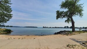 A lakeside view with a concrete walkway, trees, and a distant shoreline. - Sodus Bay View at the Point (Sodus Point)