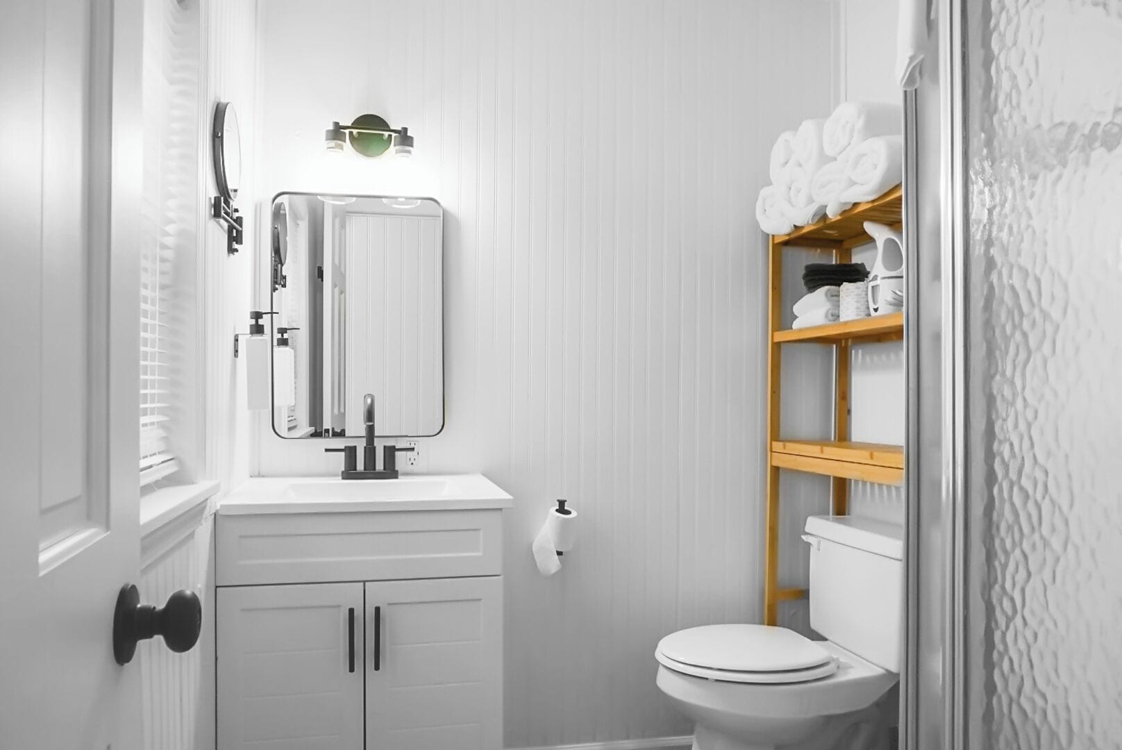 Un baño con lavamanos blanco, espejo y retrete. Hay un toallero con toallas dobladas sobre el inodoro.