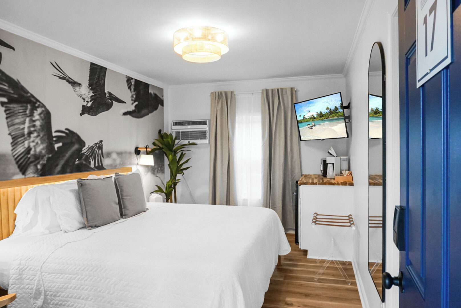 A hotel room with a bed, a TV on the wall, a desk, and a view of a beach through the window.