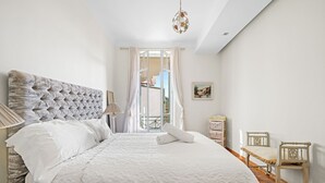 Un dormitorio con cabecera capitonada, una cama grande con sábanas blancas, una mesita de noche de madera con lámpara, una silla de madera y vistas al exterior a través de una ventana con cortinas.