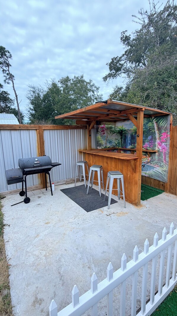 Un patio trasero con zona de barbacoa, una pérgola de madera y cerca de piquetes blancos.