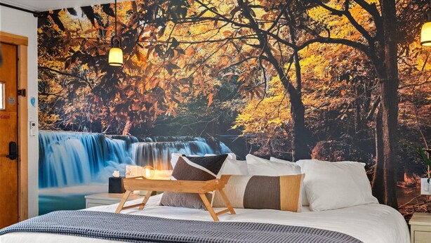 Een slaapkamer met een groot muurdecoratie van een waterval en herfstbladeren, een bed met witte beddengoed, en een nachtkastje van hout.