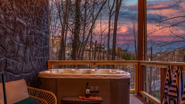 A hot tub on a balcony with a stone wall, a wicker sofa, and a table with wine glasses and a bottle. - Freshly Renovated! Pickleball, Hot Tub, Game Rm! (Gatlinburg)