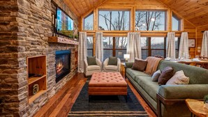 A cozy cabin interior with a stone fireplace, wooden ceiling, and large windows offering a view of the outdoors. - Freshly Renovated! Pickleball, Hot Tub, Game Rm! (Gatlinburg)