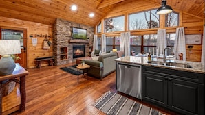 A cozy cabin interior with a stone fireplace, wooden floors, a kitchen with granite countertops, and large windows. - Freshly Renovated! Pickleball, Hot Tub, Game Rm! (Gatlinburg)