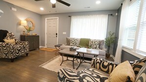 A living room with a sofa, armchair, and coffee table. There is a door, a window with curtains, and a ceiling fan. - 3-bedroom 2 full bath bungalow in the heart of the city minutes from DT Raleigh. (Raleigh)