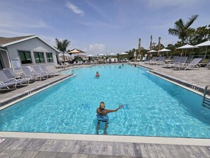 Une piscine entourée de chaises longues et de parasols, bordée de palmiers et d’un bâtiment doté de grandes fenêtres.