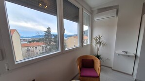A room with two wicker chairs, a small table with a vase, and a view of a cityscape through large windows. - Mystic Grey Studio Apartment (Ploce)
