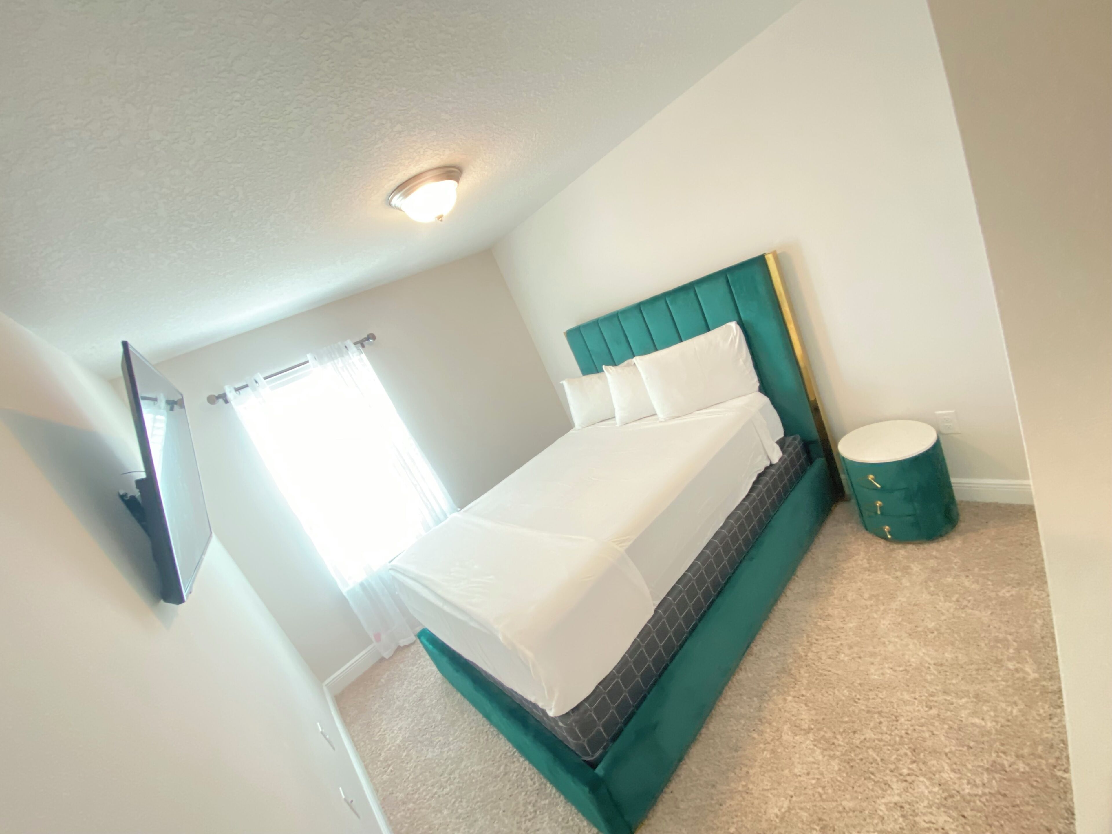Een slaapkamer met een bed, een plafondlamp, een raam met gordijnen, en een groen cilindervormig voorwerp.