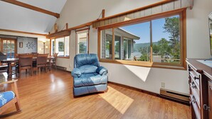 A spacious room with a large window, wooden flooring, a blue leather chair, and a dining area with a table and chairs. - Private Dock & Lake Views: Home on Lime Lake (Machias)