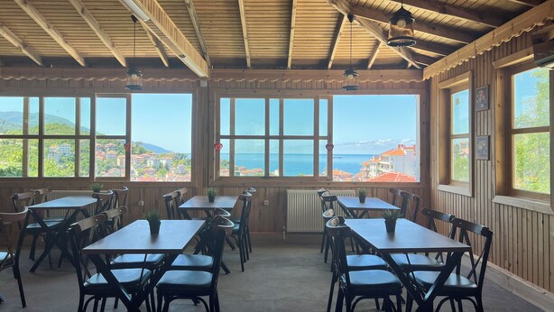 A dining area with wooden tables and chairs, large windows with a view of the sea and buildings, and a wooden ceiling with exposed beams. - İnebolu EsKonak Otel (Inebolu)