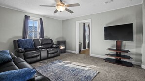 A living room with a black leather sofa, a flat-screen TV on a wooden stand, a ceiling fan, and a fireplace.