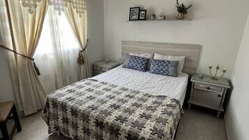 Un dormitorio con cama, mesitas de noche, una silla y una ventana con cortinas.