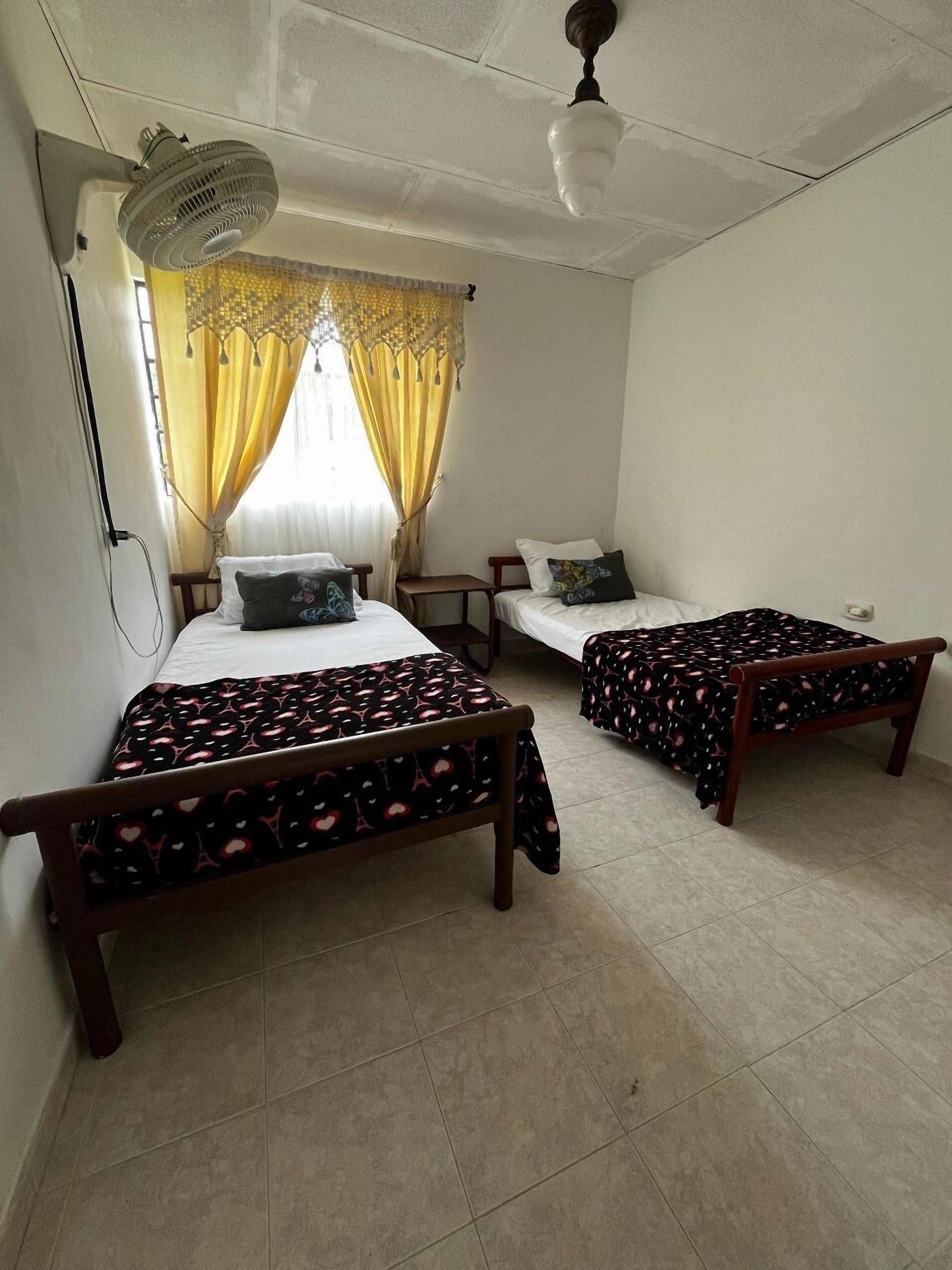 Une chambre avec deux lits en bois, un ventilateur de plafond et une fenêtre avec des rideaux jaunes.
