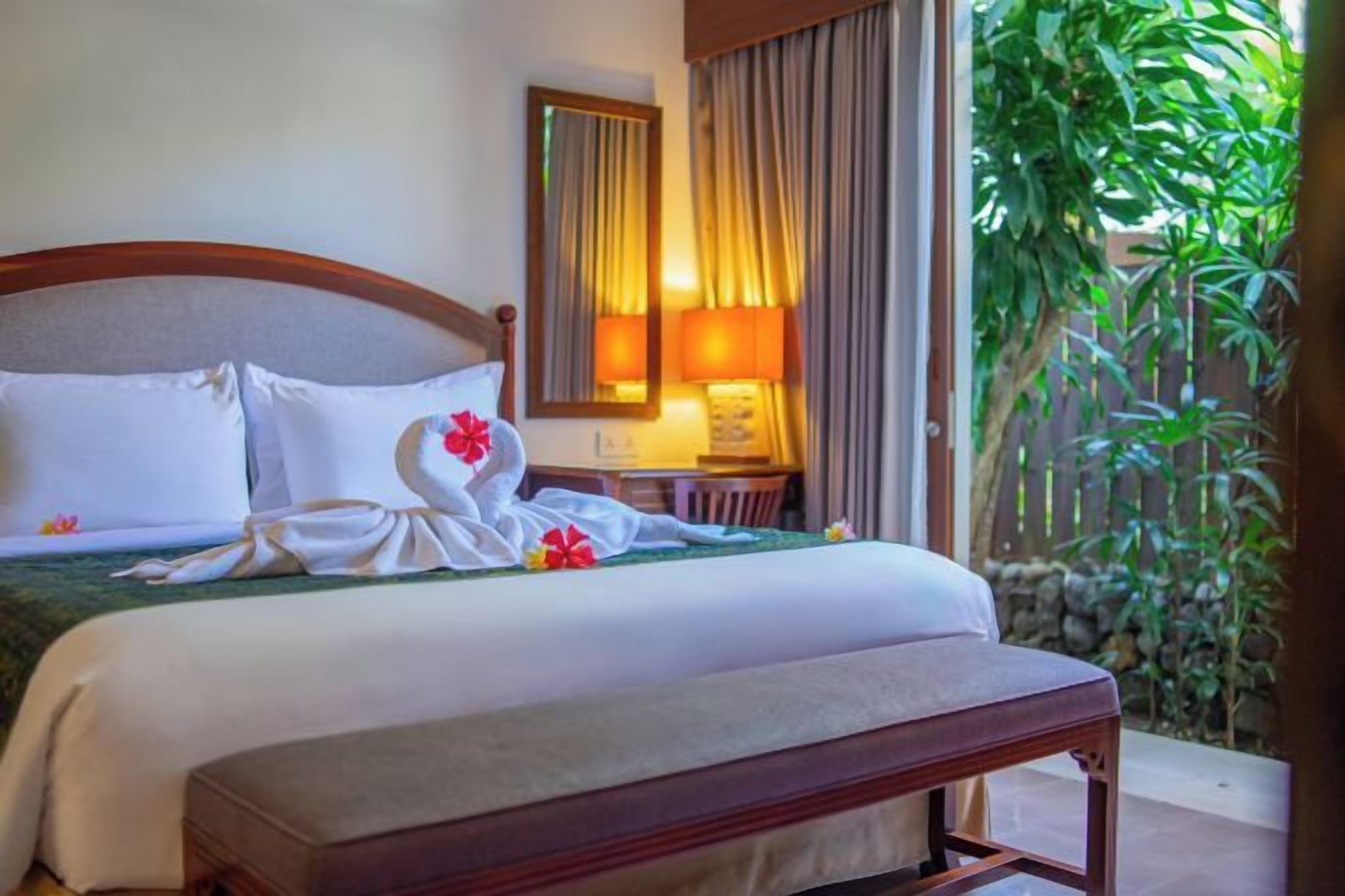 A hotel room with a bed, a bedside table, a lamp, a mirror, and a view of greenery.