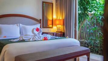 A hotel room with a bed, a bedside table, a lamp, a mirror, and a view of greenery.