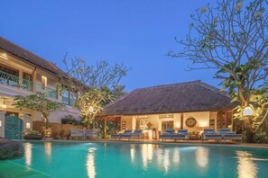 A pool area with lounge chairs, a thatched-roof structure, and a building with balconies. - Kama Village Sanur (Bali)
