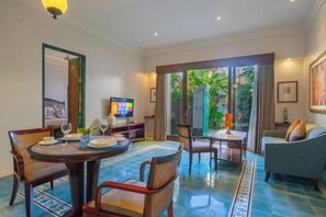 Quarto de hotel com área de jantar, sofá, TV e vista para o verde pelas janelas.
