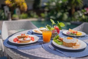 A round table with breakfast plates, a glass of orange juice, and a pool in the background. - Kama Village Sanur (Bali)