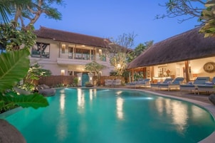 A pool with lounge chairs, a building with a balcony, and a thatched-roof structure.