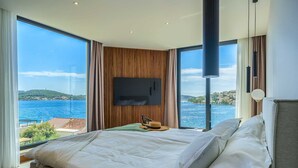 A modern bedroom with a large bed, a flat-screen TV mounted on the wall, and a view of the sea. - Luxurious 5-BR Villa for 10 with Panoramic Views and Heated Pool (Zatoglav)