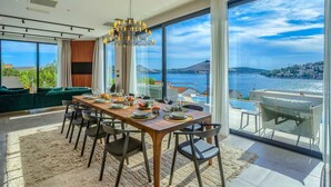 A modern dining room with a large table set for six, large windows offering a view of the sea and a cityscape, and a stylish chandelier. - Luxurious 5-BR Villa for 10 with Panoramic Views and Heated Pool (Zatoglav)