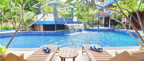 A pool with a waterfall feature, surrounded by wooden lounge chairs and a small table.