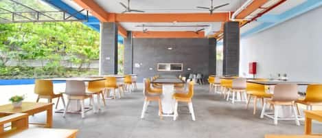 A modern indoor space with tables and chairs arranged for dining, a kitchen area in the background, and a view of greenery outside.