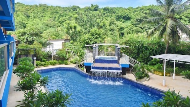 A swimming pool with a waterfall feature, surrounded by lush greenery and a mountainous backdrop.