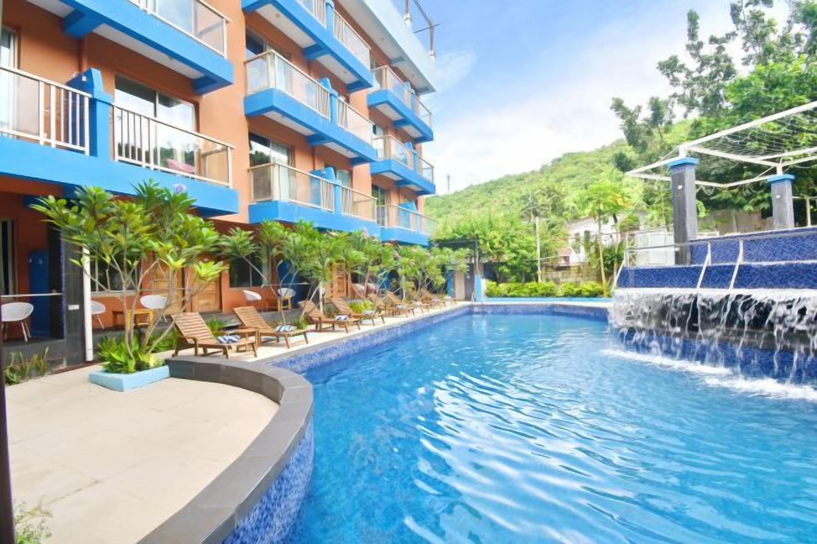 A hotel pool area with a clear blue pool, a waterfall feature, and a building with balconies.