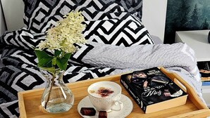 A bed with a geometric patterned bedspread, a bedside table with a tray holding a book, a cup of coffee, and a vase with flowers.