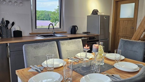 A dining area with a wooden table set for four, a kitchen with a refrigerator, and a window with a view of trees and flowers.