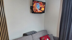 A grey sofa with two red pillows, a flat-screen TV mounted on the wall, and a wooden dining table with chairs.