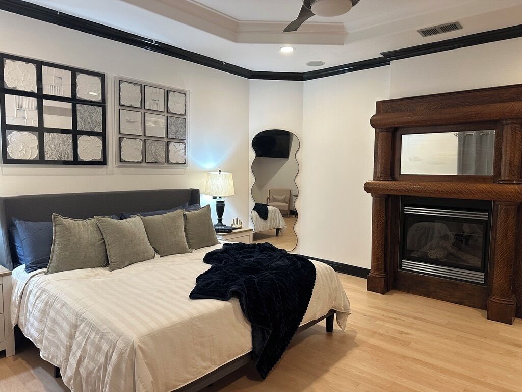 A bedroom with a bed, bedside tables, a lamp, a mirror, a fireplace, and artwork on the wall.