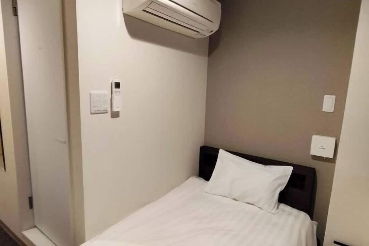 A single bed with a white mattress and pillow, a wall-mounted air conditioner, and a light switch on the wall.