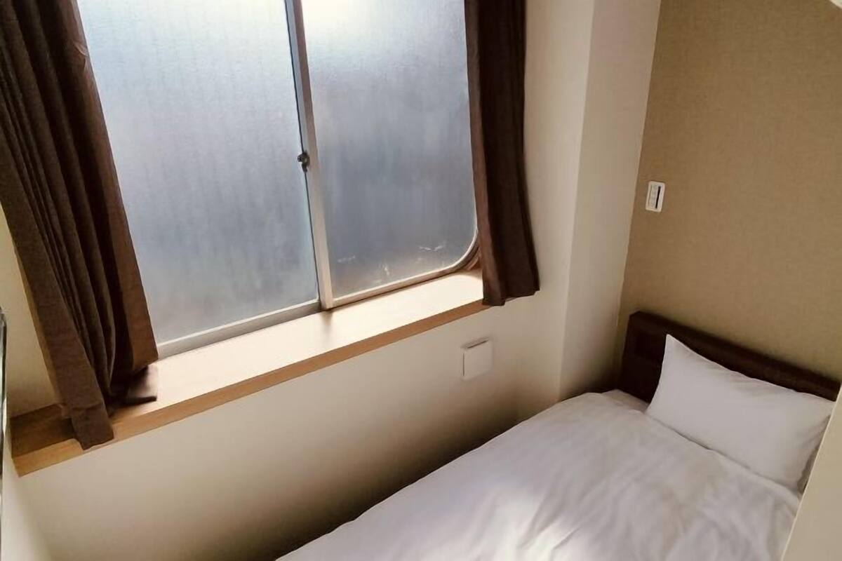 A small, single-bed room with a window, a wooden shelf, and a wall socket.