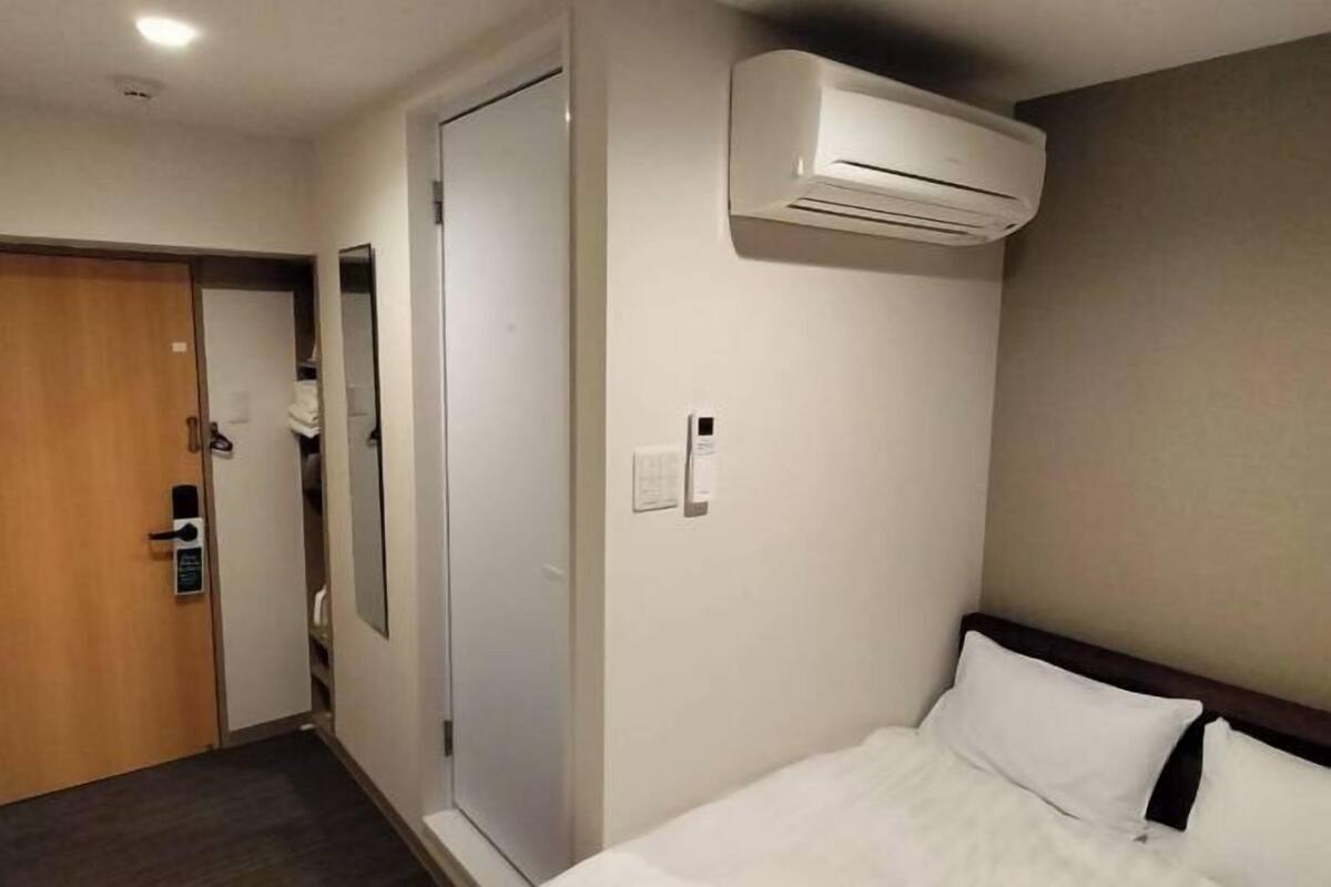 A hotel room with a bed, an air conditioning unit, a door with a handle, and a wall-mounted light switch.