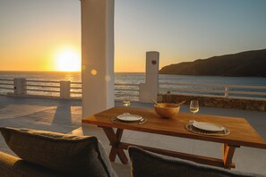 A wooden table with a white plate, a bowl, and a wine glass set for two, overlooking a coastal sunset.