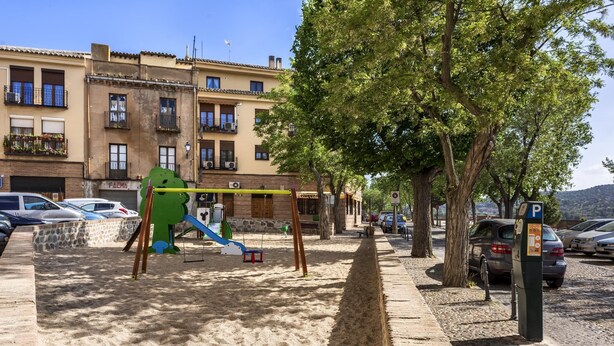 Un parque infantil con columpios y toboganes, un estacionamiento con coches, y un edificio con balcones al fondo.