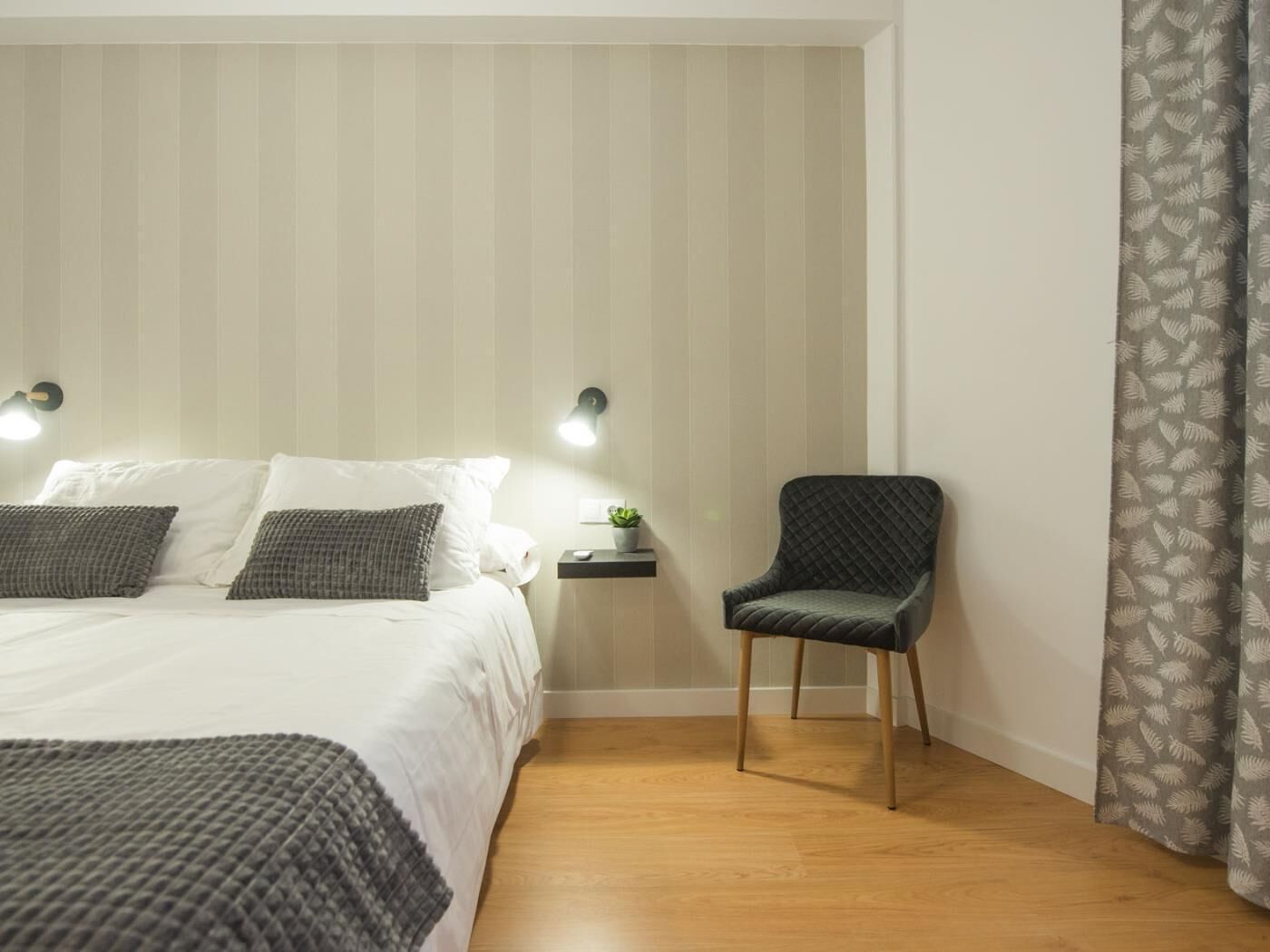 Een slaapkamer met een bed, bedlampjes, een stoel en een nachtkastje met een plant.