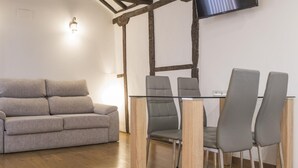 A modern living room with a grey sofa, a glass-top dining table with four grey chairs, wooden beams on the ceiling, and a flat-screen TV mounted on the wall.