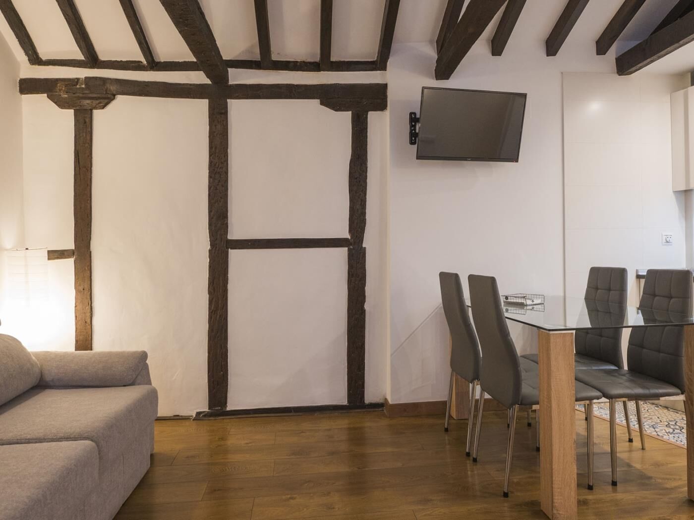 Een kamer met zichtbare houten balken, een grijze bank, een glazen tafel met vier stoelen en een flatscreen televisie aan de muur.