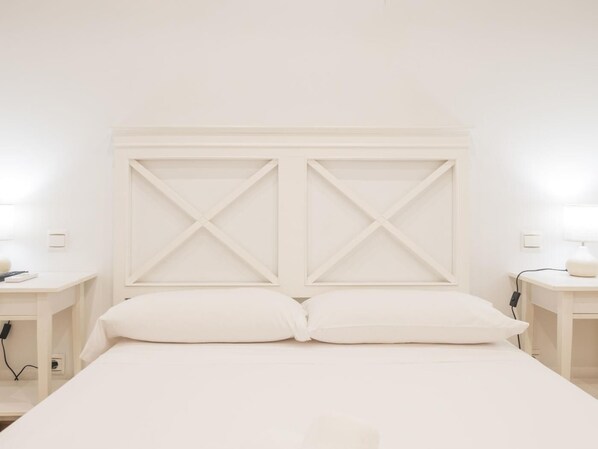 A minimalist white bedroom with a large bed, two nightstands, and a simple headboard.