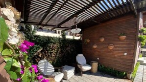 En udendørs patio med flisebelagt gulv, en træpergola og siddepladser bestående af en sofa og stole.
