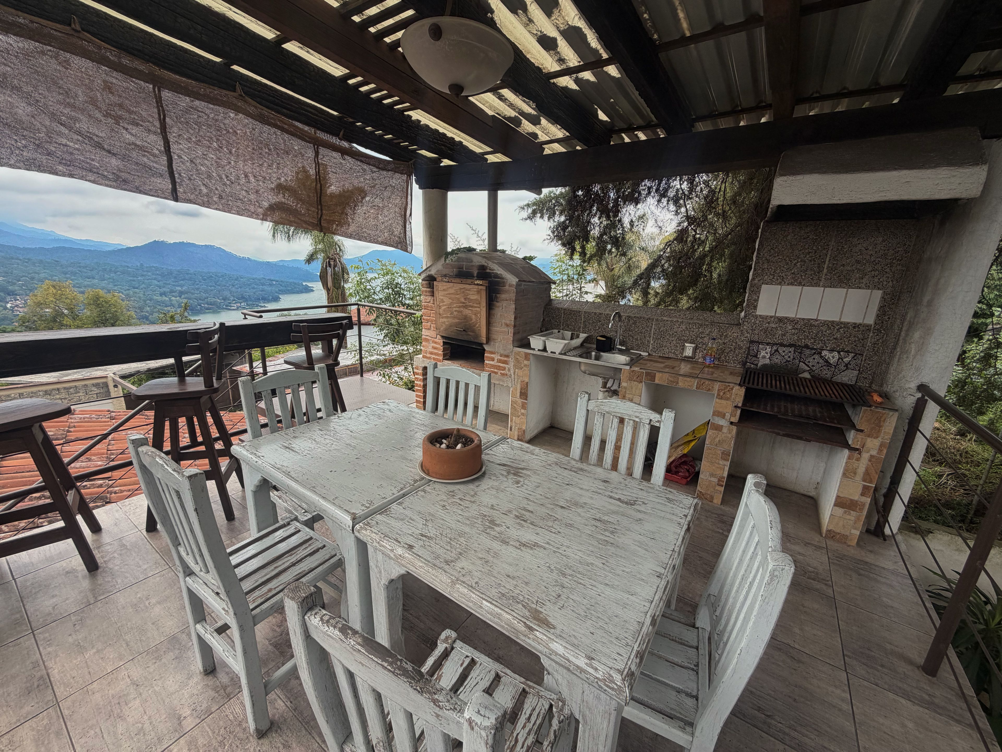 An outdoor patio with a rustic wooden table and chairs, a brick oven, and a view of the mountains.