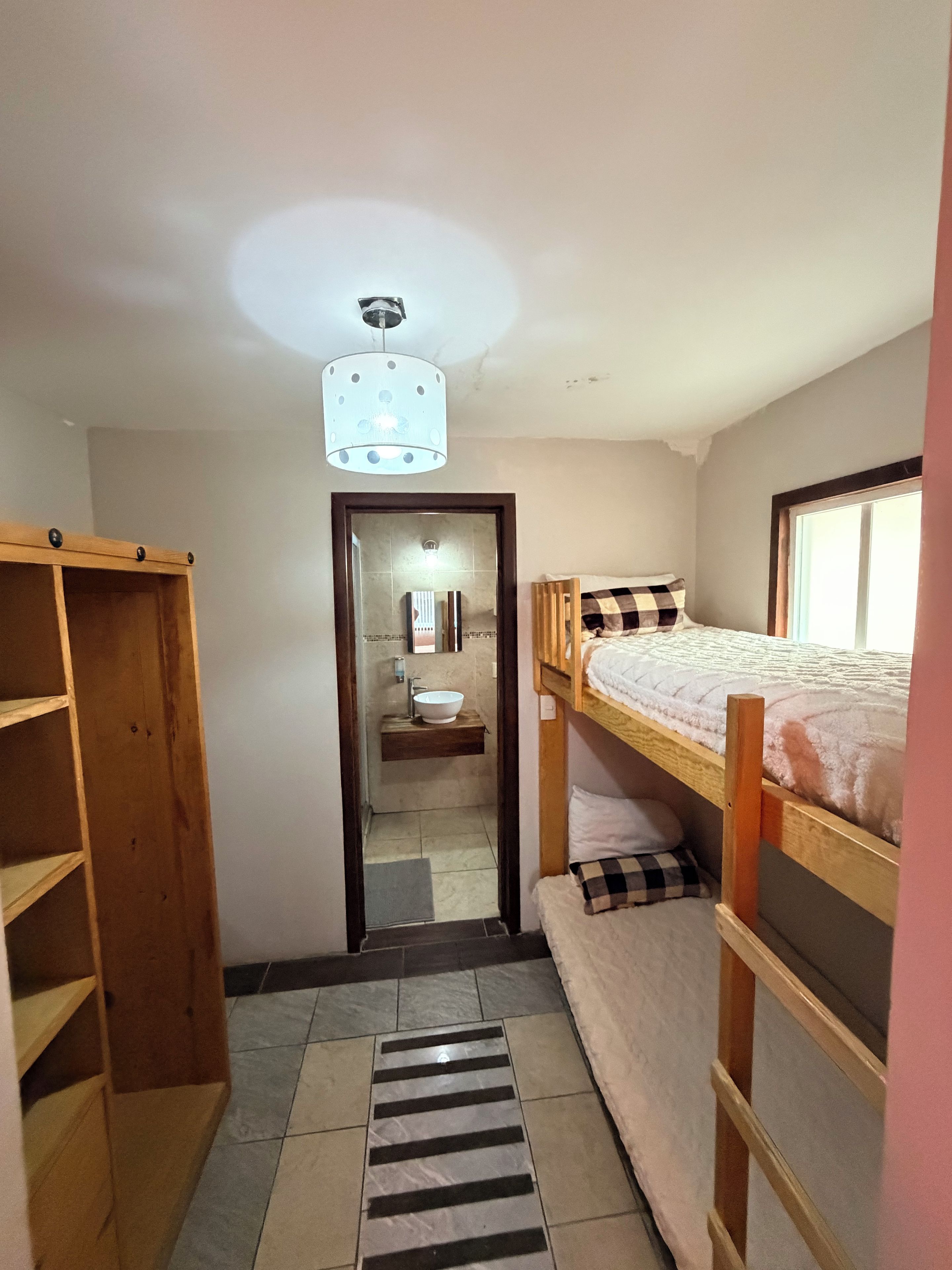 A compact room with a bunk bed, a wooden wardrobe, a bathroom with a mirror and sink, and a striped mat on the floor.
